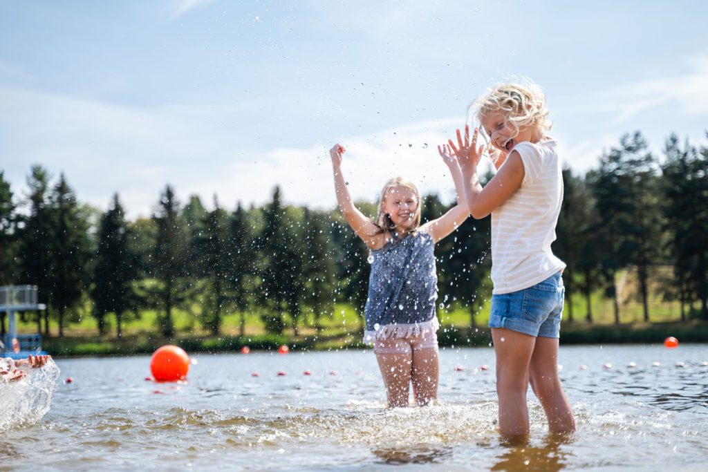 Zwei fröhliche Kinder spielen im seichten Wasser eines Badesees, spritzen Wasser in die Luft; im Hintergrund Wald und Bojen im See. Foto: Oliver Göhler