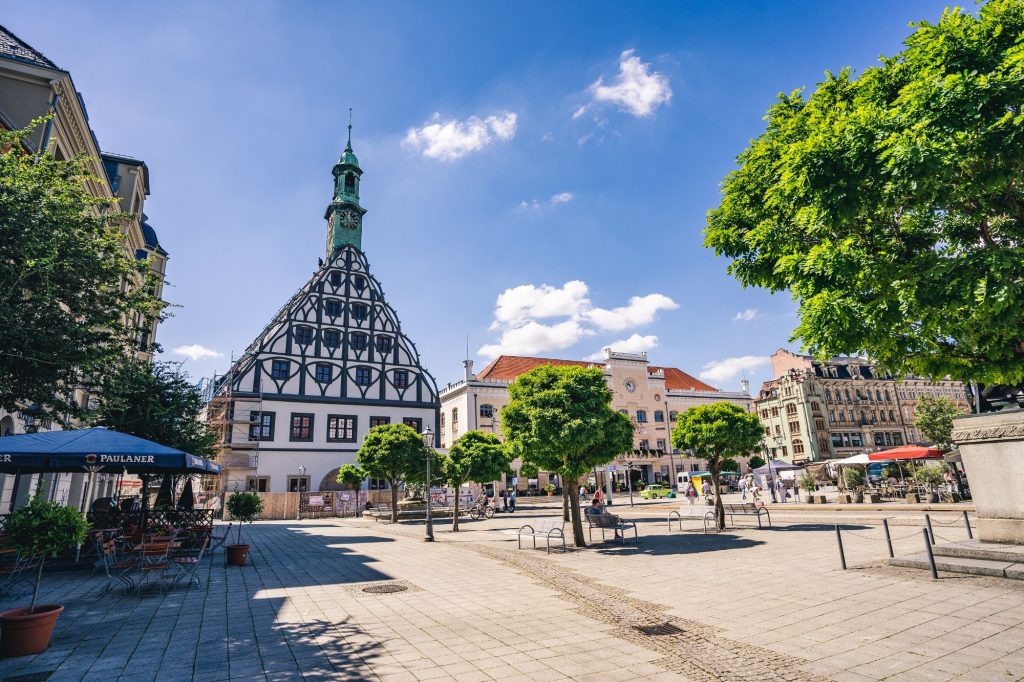Historisches Gewandhaus mit Fachwerkfassade und spitzem Turm auf dem Hauptmarkt von Zwickau, umgeben von weiteren Altbauten und Bäumen.