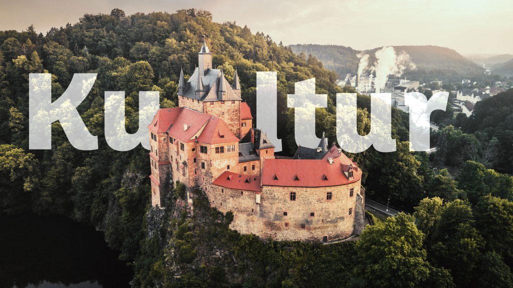 Luftaufnahme der mittelalterlichen Burg Kriebstein auf einem Felssporn über dem Fluss, umgeben von dichtem Wald; im Bild groß der Schriftzug ‚Kultur‘.