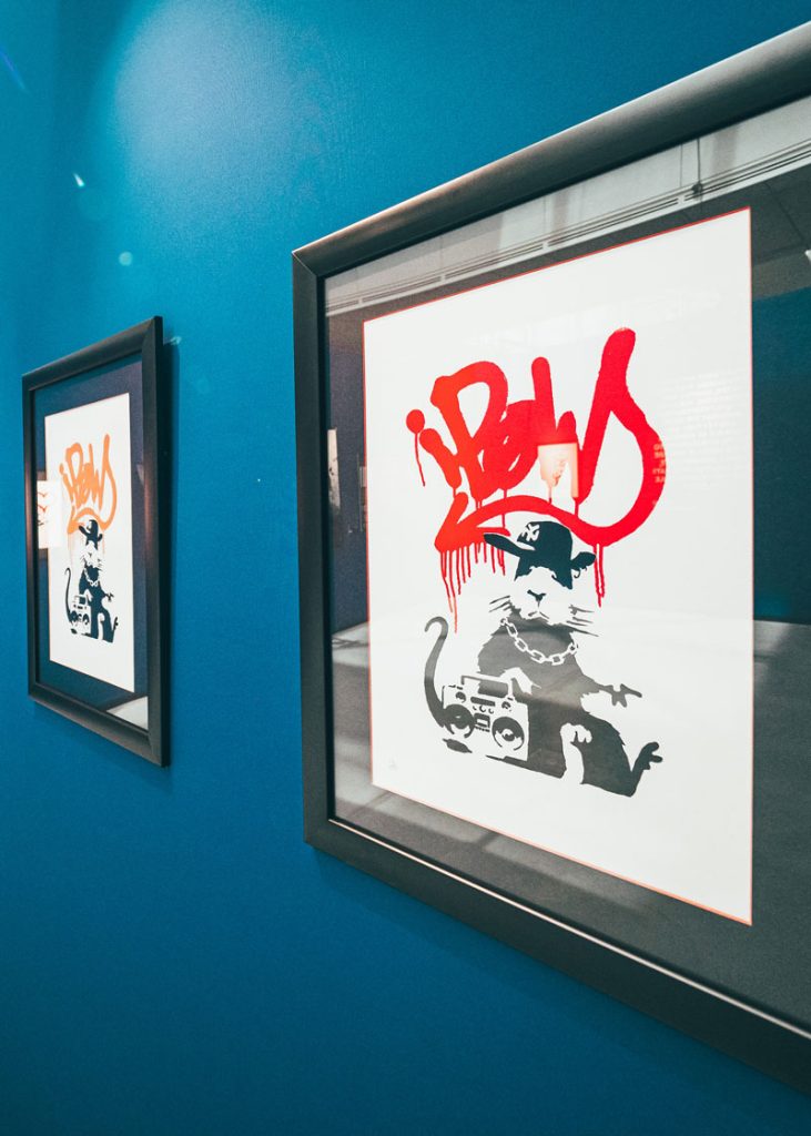Zwei gerahmte Drucke einer graffitiartigen Ratte mit rotem Schriftzug an einer blauen Ausstellungswand – Kunstwerke im Stil von Banksy.