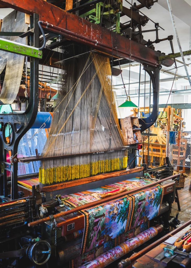 Historischer Jacquard-Webstuhl mit buntem Stoff in einer alten Textilfabrik, im Hintergrund weitere Webmaschinen und Werkstatt-Atmosphäre.