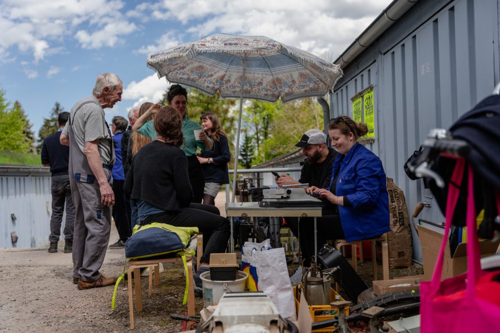 Mehrere Menschen treffen sich draußen an einem Tisch mit alten Geräten und Werkzeugen. Zwei Personen sitzen unter einem Sonnenschirm und tippen auf einer Schreibmaschine. Andere stehen drumherum, unterhalten sich, begutachten die Gegenstände und lachen. Es wirkt wie ein Flohmarkt.