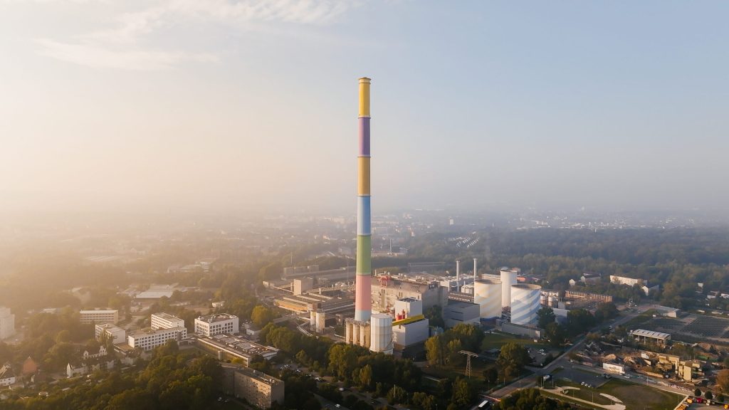 Eine Luftaufnahme von einem sehr hoher Schornstein mit bunten Farbringen steht in einem Industriegebiet in der Stadt Chemnitz. Um den Schornstein herum sieht man viele Gebäude, Straßen und Bäume. Der Himmel ist hell, und über der Stadt liegt leichter Dunst.