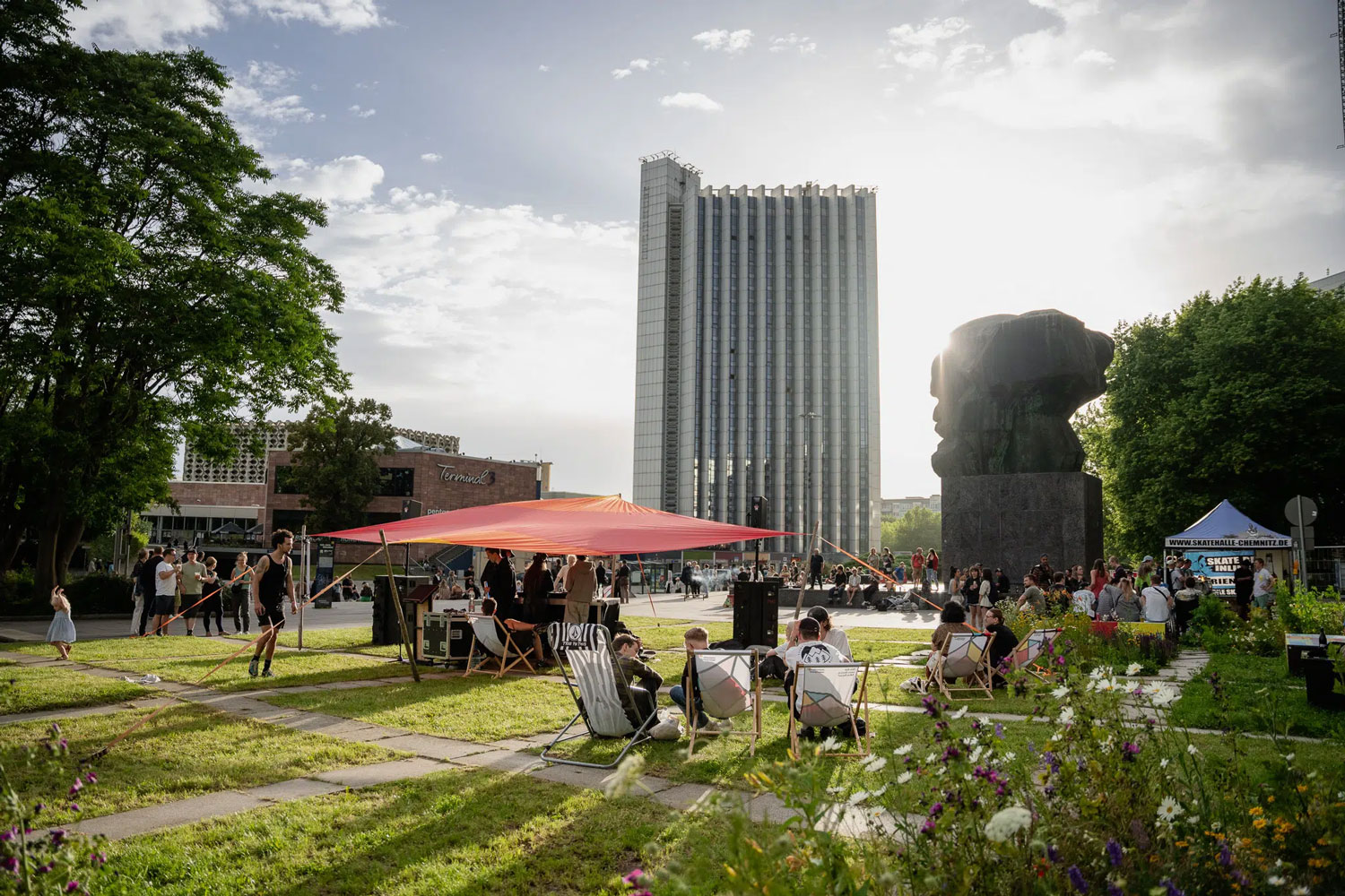 Menschen sitzen auf Liegestühlen und unter einem Sonnensegel auf einer Wiese vor dem Karl-Marx-Monument in Chemnitz. Die Nachmittagssonne hüllt das Geschehen in ein warmes Licht und betont die Statur. Im Hintergrund stehen hohe Gebäude, Blumen blühen im Vordergrund, und es herrscht eine entspannte, sommerliche Stimmung.