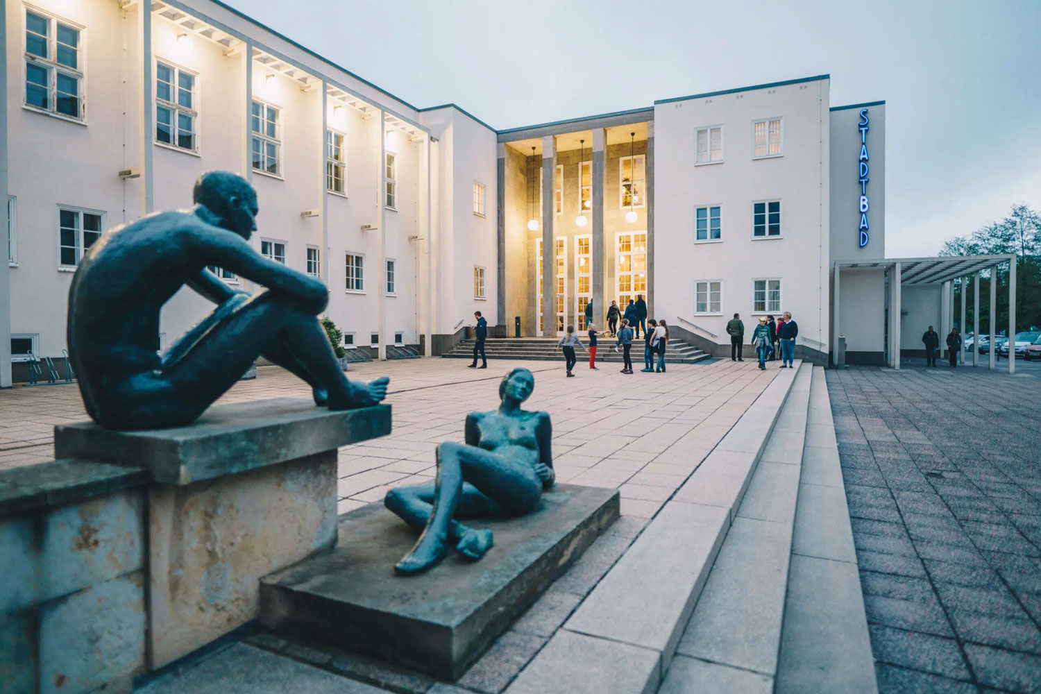 Ein weißes Schwimmbad-Gebäude mit vielen Fenstern. Vor dem Eingang stehen zwei große Figuren aus Bronze. Die Figuren zeigen nackte Menschen, die sitzen. Am Eingang gehen Menschen eine Treppe hoch. Es ist Abend und das Licht im Gebäude leuchtet.