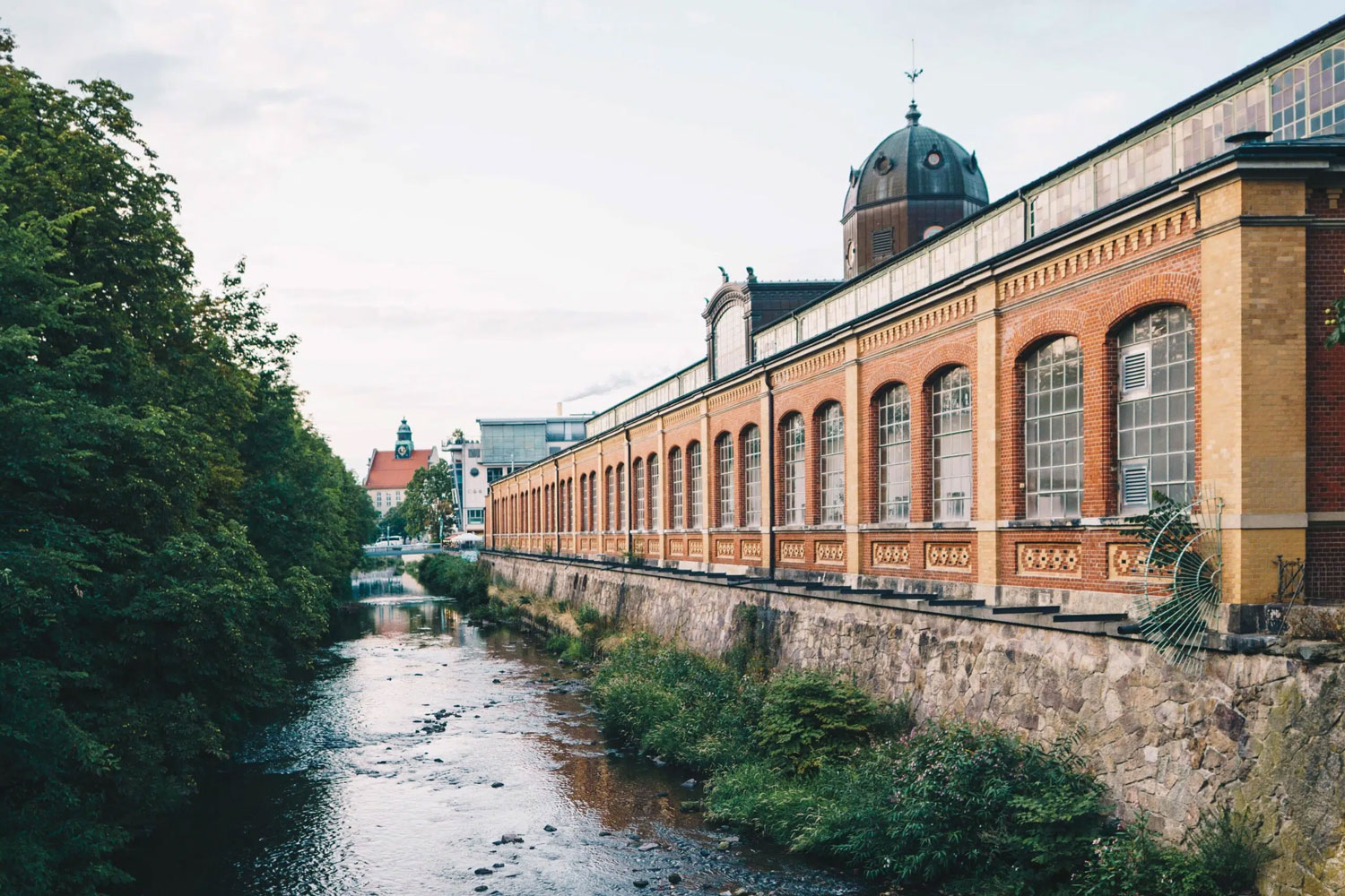 in langes Backsteingebäude mit großen Fenstern steht direkt an einem Fluss. Links wachsen viele Bäume am Ufer, rechts zieht sich das Gebäude mit einem kleinen Turm in die Tiefe. Im Hintergrund sieht man die Dächer der Stadt.