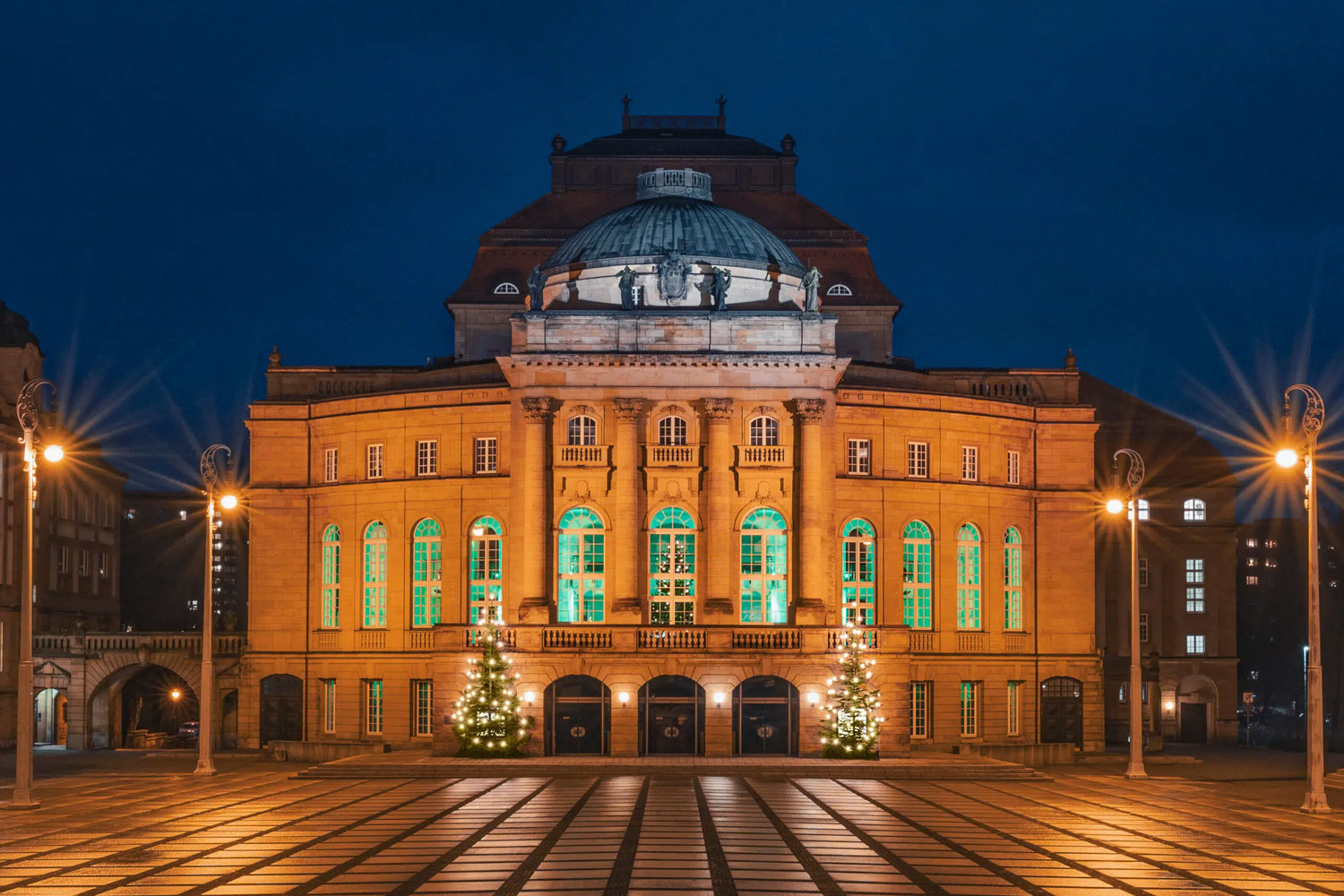 Opernhaus am Theaterplatz am Abend mit beleuchteten Fenstern. Davor stehen zwei beleuchtete Weihnachtsbäume Foto: Theater Chemnitz