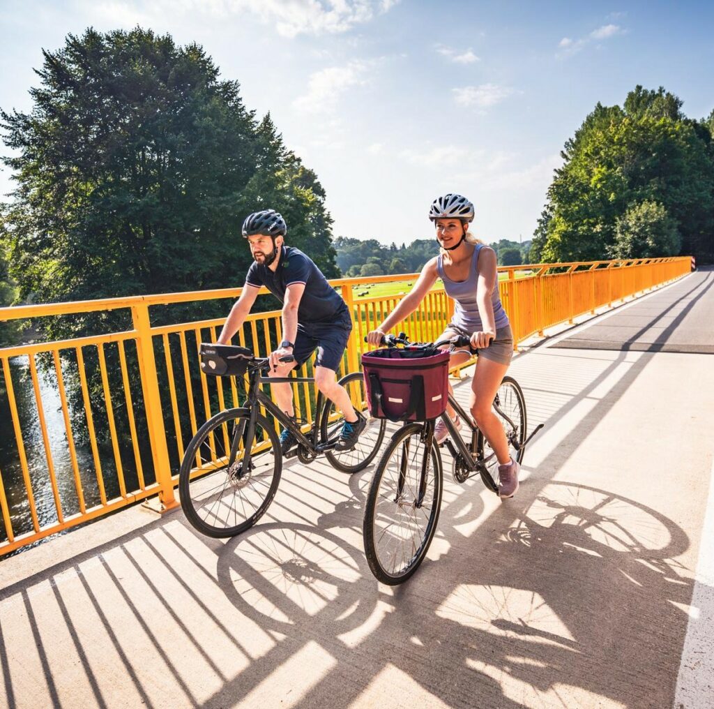 Zwei Radfahrer überqueren bei Sonnenschein eine gelbe Brücke auf dem Chemnitztalradweg, umgeben von Natur. Foto: Zweckverband "Chemnitztalradweg", Oliver Göhler