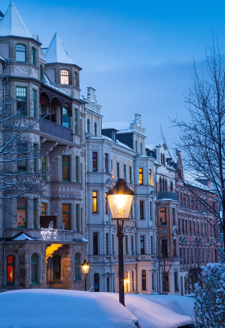 Eine Reihe historischer Villen mit Türmchen, Erkern und reich verzierten Fassaden säumt eine verschneite Straße. Warm beleuchtete Fenster und Straßenlaternen setzen stimmungsvolle Akzente in der blauen Abenddämmerung. Kahle Bäume und schneebedeckte Autos unterstreichen die ruhige winterliche Atmosphäre. Foto: Dirk Hanus