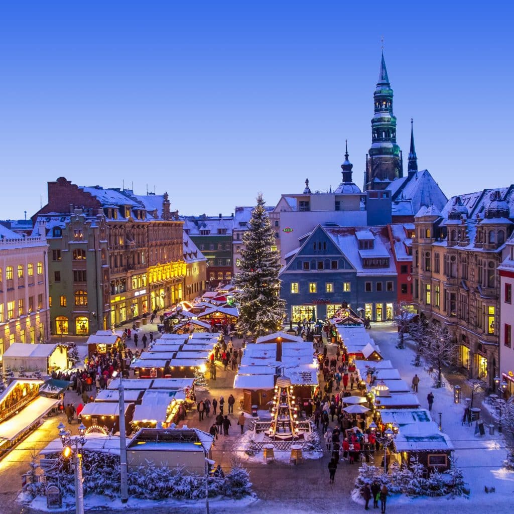 Der verschneite Weihnachtsmarkt erstreckt sich über den historischen Hauptmarkt von Zwickau. Zahlreiche beleuchtete Verkaufsstände und ein großer Weihnachtsbaum prägen den Platz, während Besucherinnen und Besucher zwischen den Buden unterwegs sind. Die umliegenden Gebäude und der Turm des Doms St. Marien bilden eine festliche winterliche Kulisse. Foto: Kultour Z_