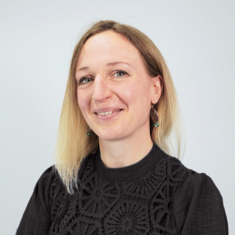 Stefanie Weise ist in einem klassischen Porträt vor einem hellen, neutralen Hintergrund zu sehen. Sie trägt ein dunkles Oberteil mit strukturierter Verzierung und blickt lächelnd in die Kamera. Ihre glatten blonden Haare fallen offen über die Schultern.