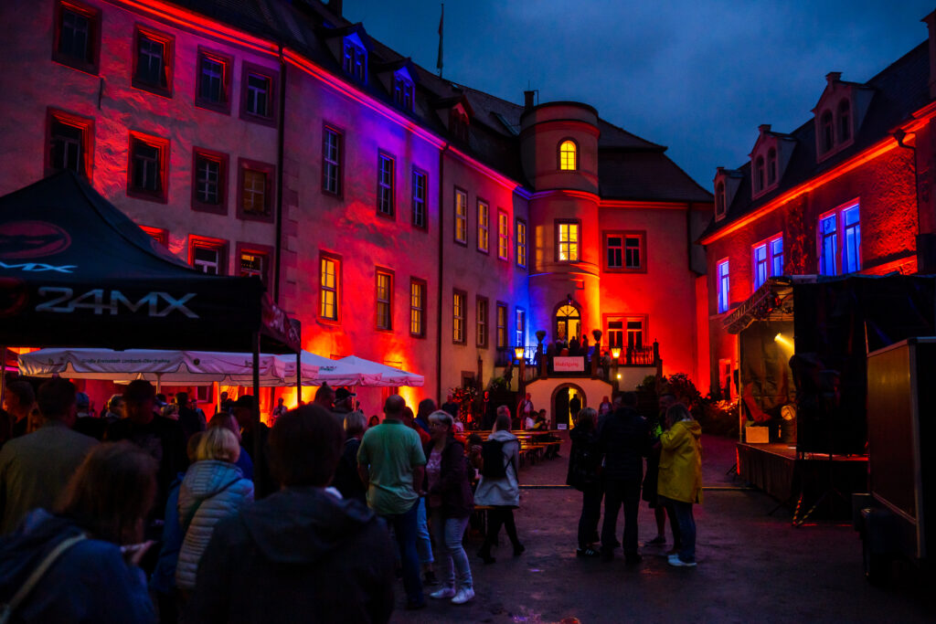 Der Innenhof einer historischen Schlossanlage, von Schloss Wolkenburg, ist am Abend in kräftiges rotes und blaues Licht getaucht. Zahlreiche Besucherinnen und Besucher stehen zwischen Zelten, Sitzgelegenheiten und einer kleinen Bühne. Beleuchtete Fenster und die farbig angestrahlten Fassaden schaffen eine lebendige Veranstaltungsatmosphäre. Foto: Oliver Göhler
